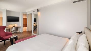 Camera d'albergo con un letto grande, una poltrona rossa, una scrivania con una sedia, una TV fissata al muro e una porta che dà su un'altra stanza.