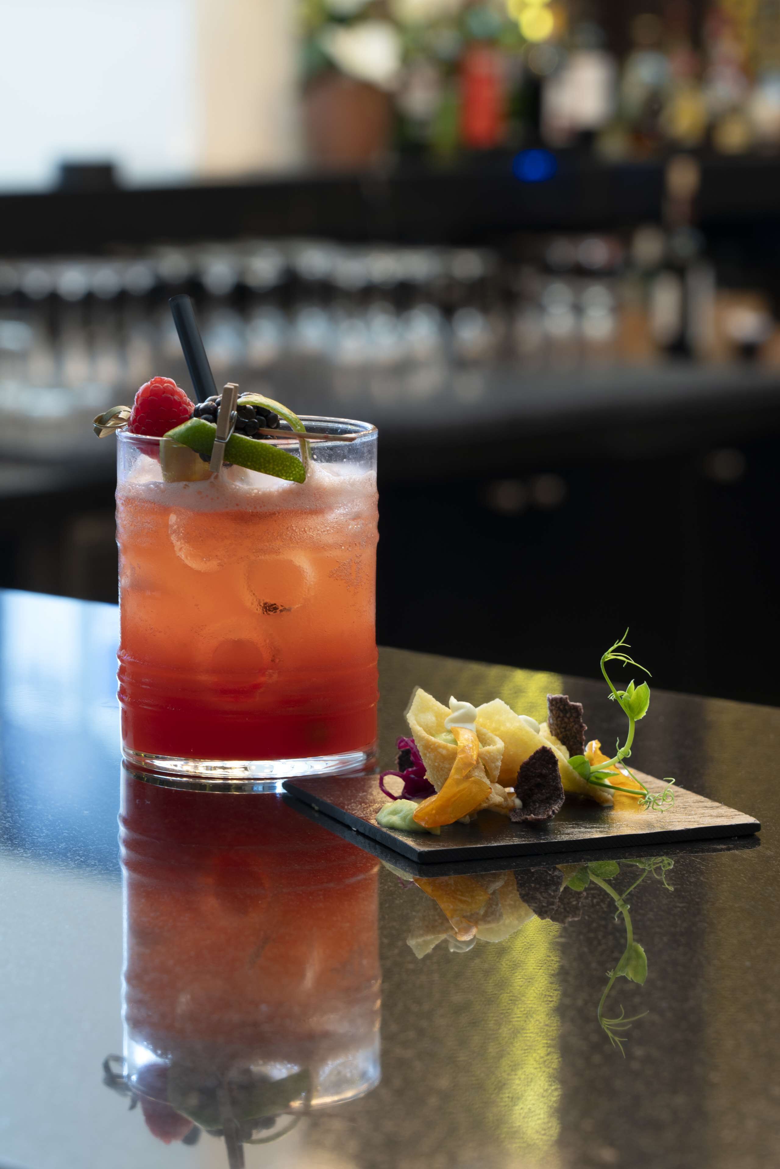 A cocktail with a garnish and a small plate of food on a reflective surface.