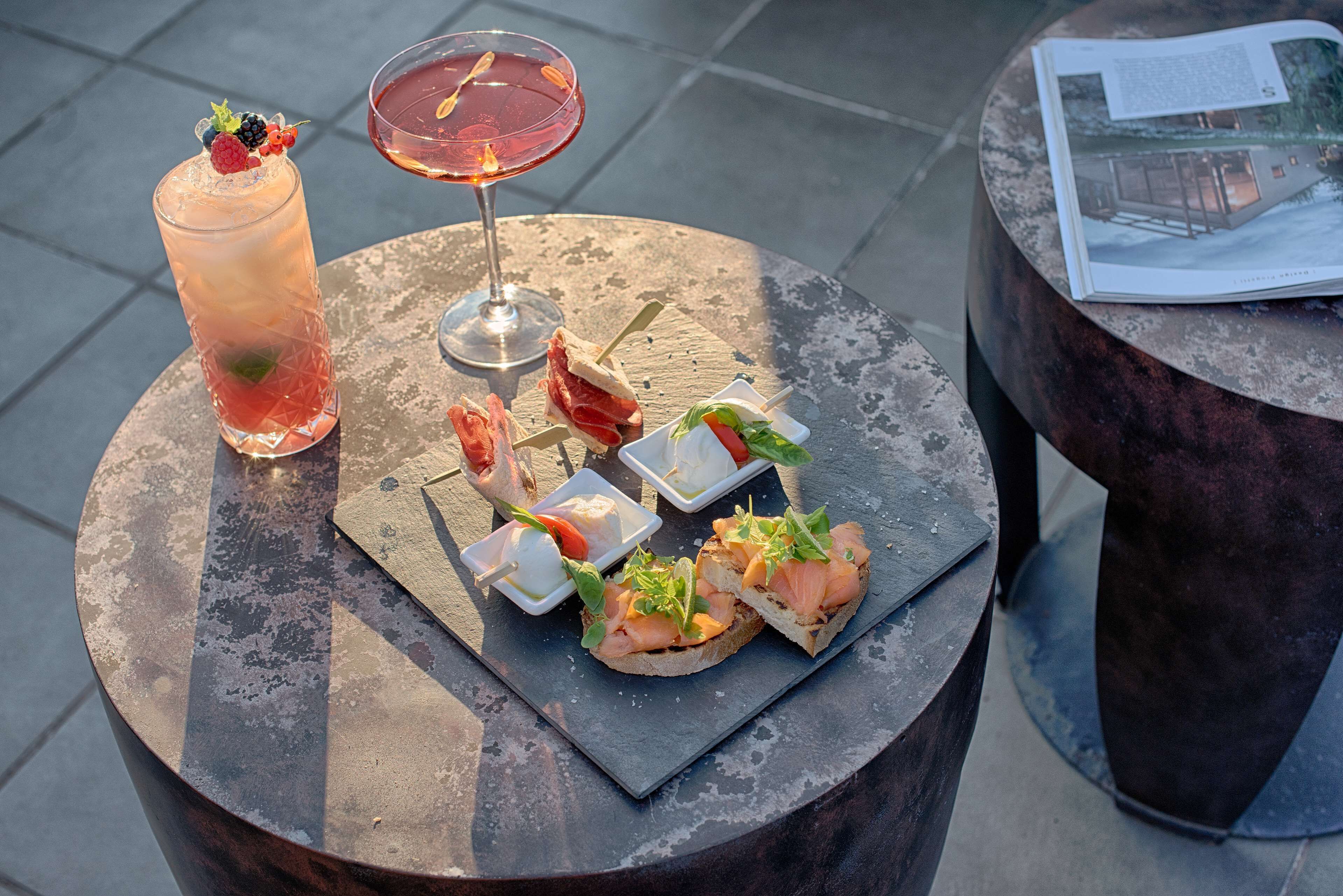 A round table with a variety of food items and drinks, including a tall glass of iced beverage, a cocktail, and several small plates with appetizers.