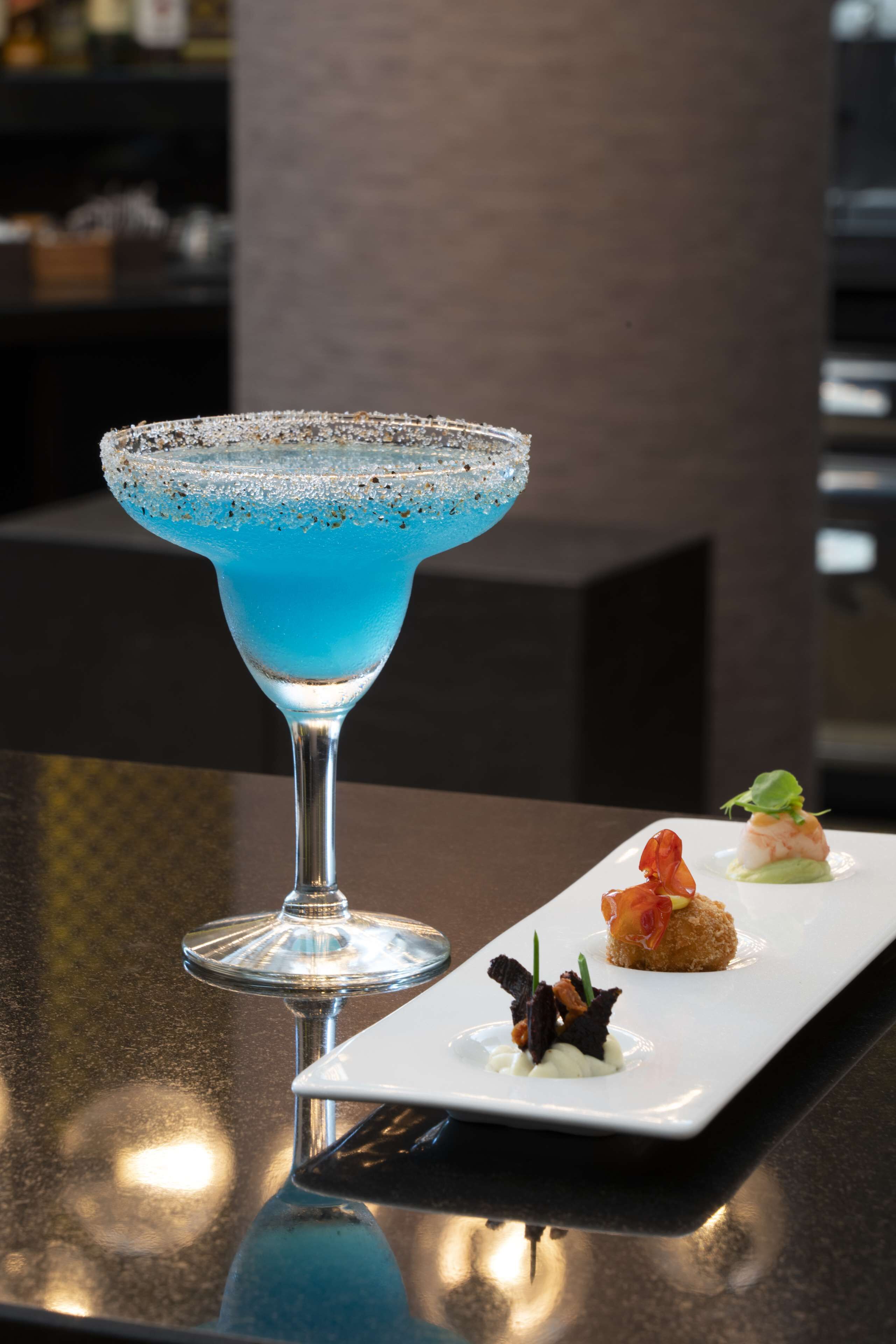 A blue cocktail in a martini glass with a rim coated in salt and pepper, accompanied by three small appetizers on a rectangular white plate.