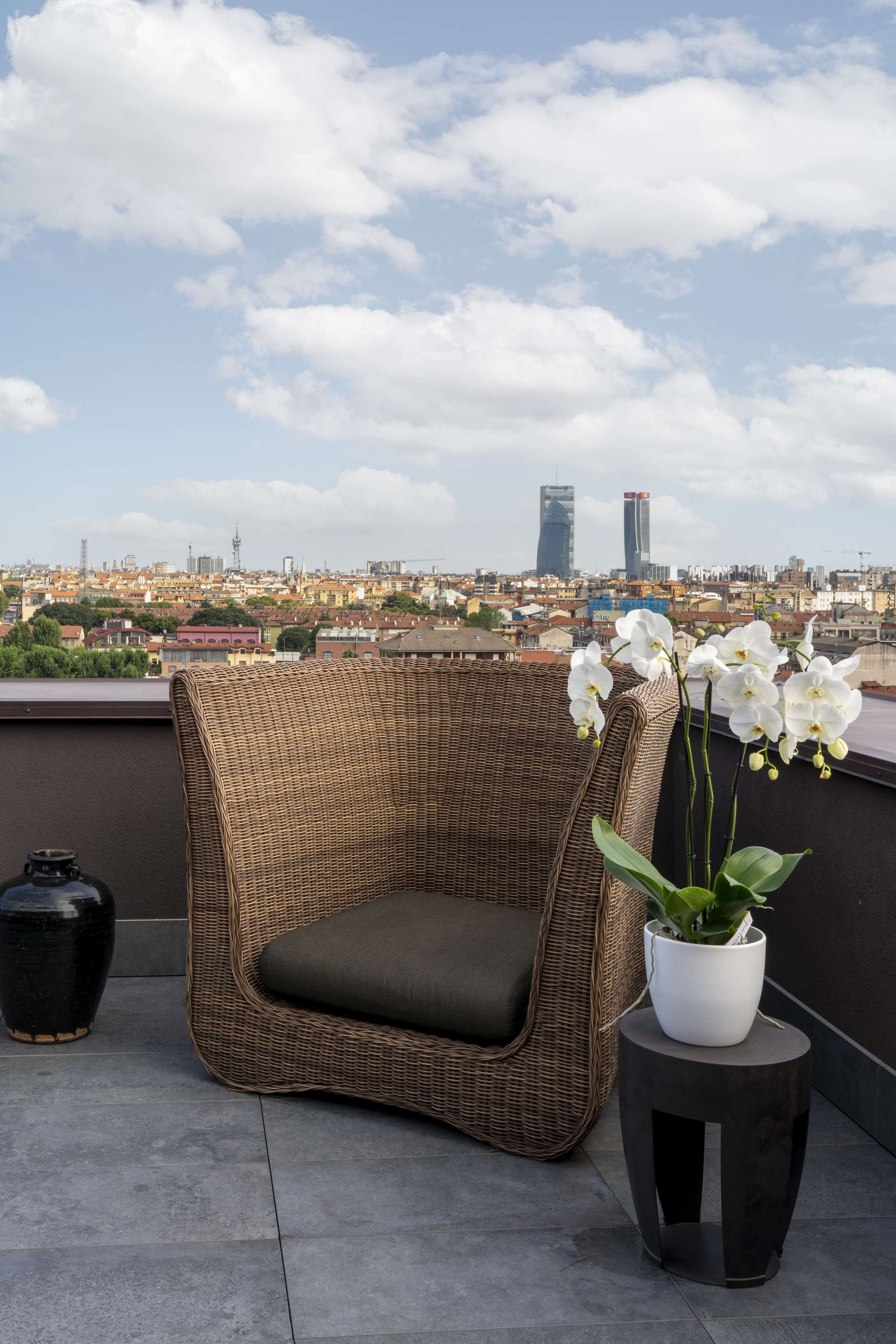Ein Korbstuhl mit Kissen auf einer Dachterrasse, ein Blumentopf und ein Blick auf eine Stadtansicht mit Gebäuden im Hintergrund.