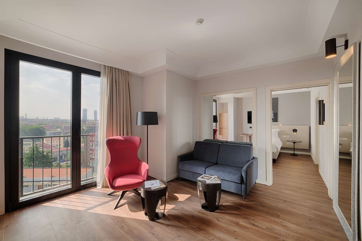 A modern living room with a large window, a red chair, a grey sofa, and a small side table.