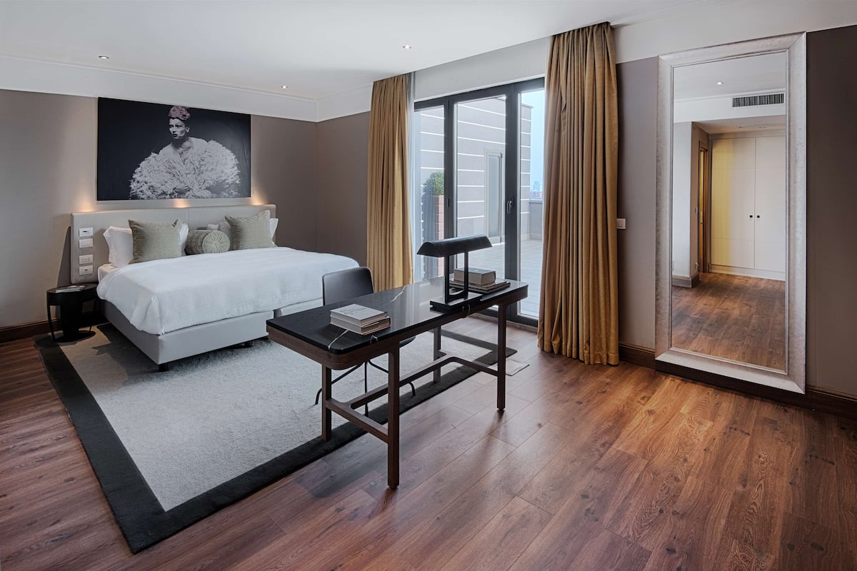 A modern hotel room with a large bed, a desk, and a view of the outdoors through a sliding door.