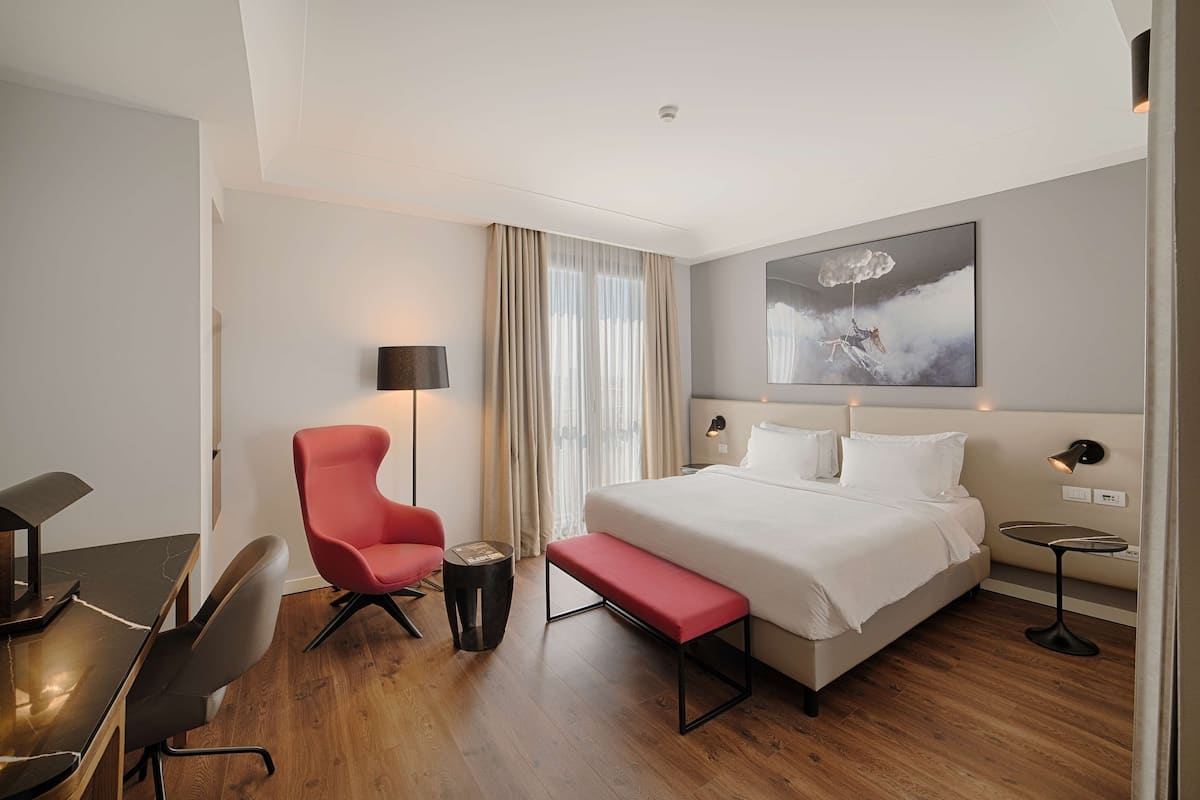 A modern hotel room with a large bed, a desk with a chair, a small side table, and a bench.