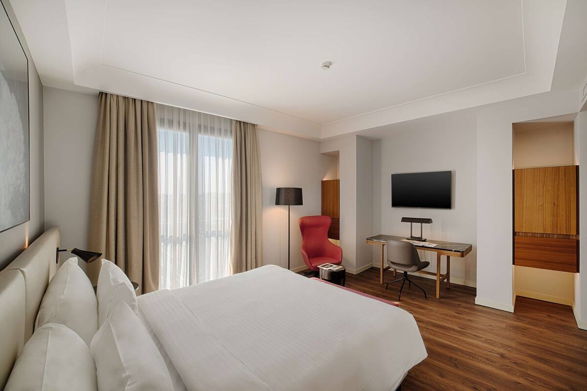 A hotel room with a large bed, a desk, a red chair, a television, and a window with curtains.