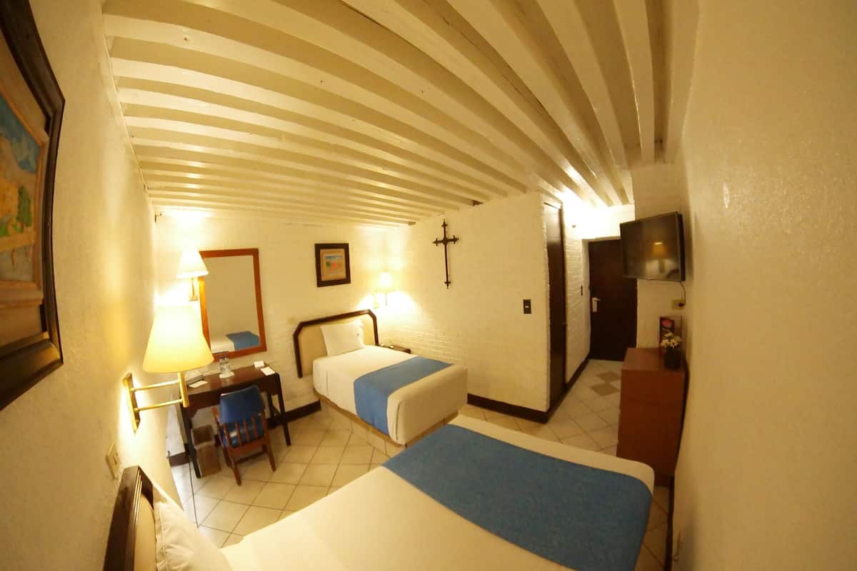 A hotel room with two beds, a desk, a chair, a lamp, and a cross on the wall.