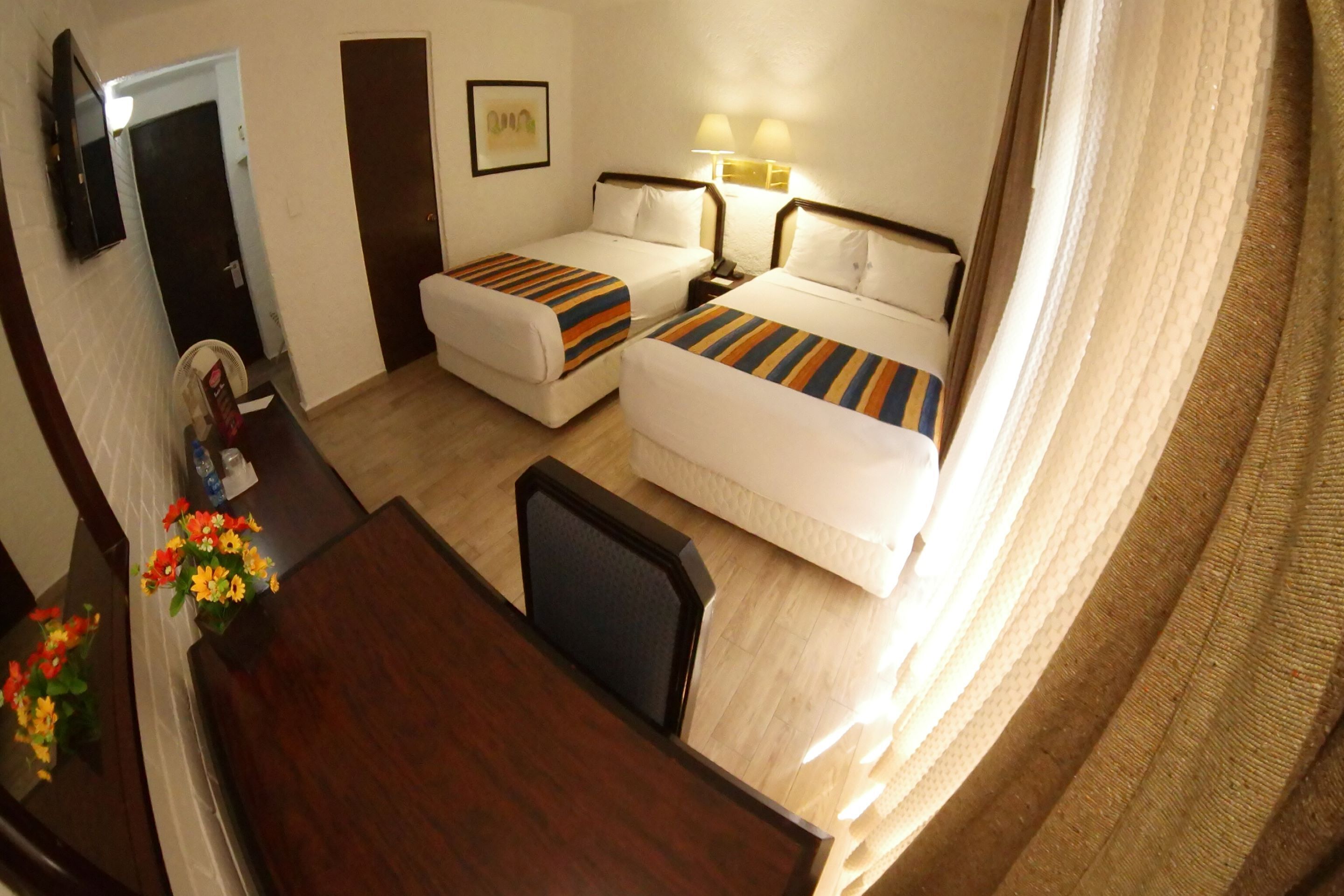 A hotel room with two beds, a desk, a chair, a TV, and a wall-mounted lamp.