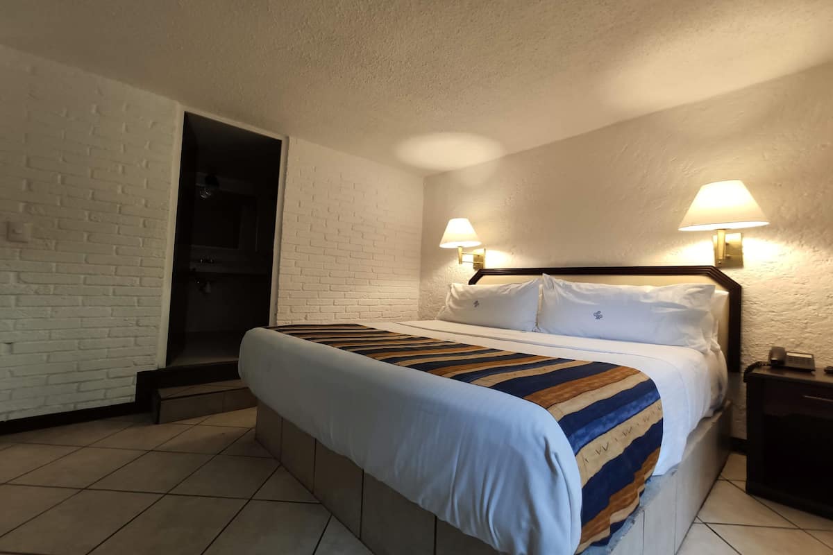 A hotel room with a bed, two bedside lamps, a white brick wall, and a door leading to a bathroom.