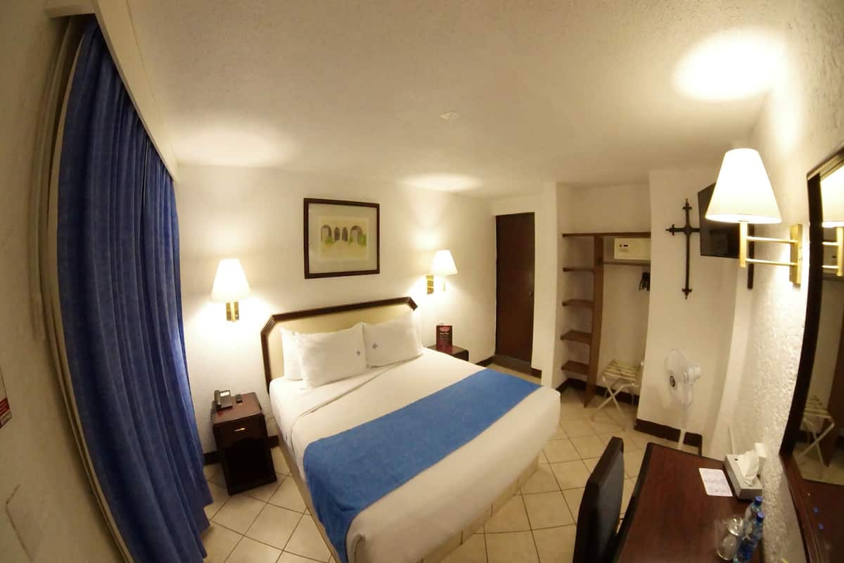 A hotel room with a bed, bedside lamps, a desk with a chair, a mirror, and a wardrobe.