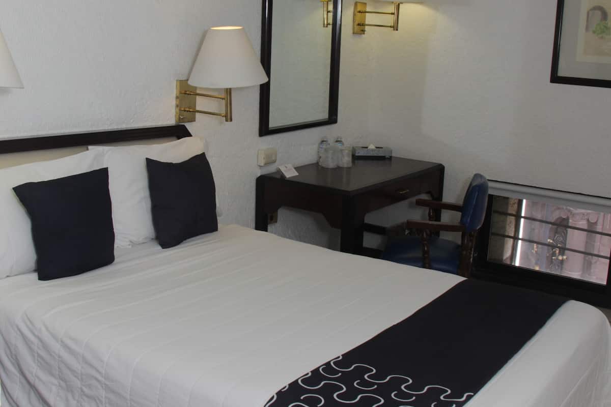 A hotel room with a bed, a desk, a chair, a lamp, a mirror, and a window.