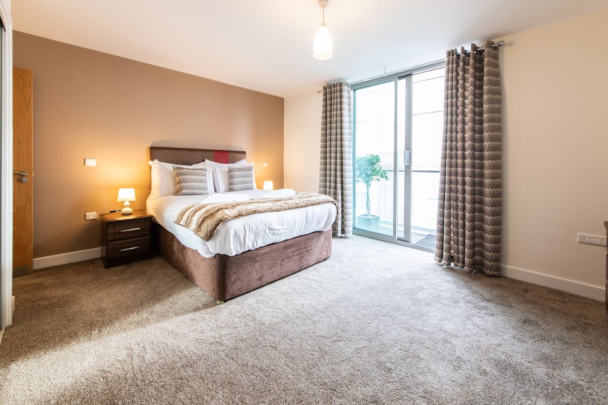 A bedroom with a large bed, a nightstand, a lamp, a dresser, and a potted plant.