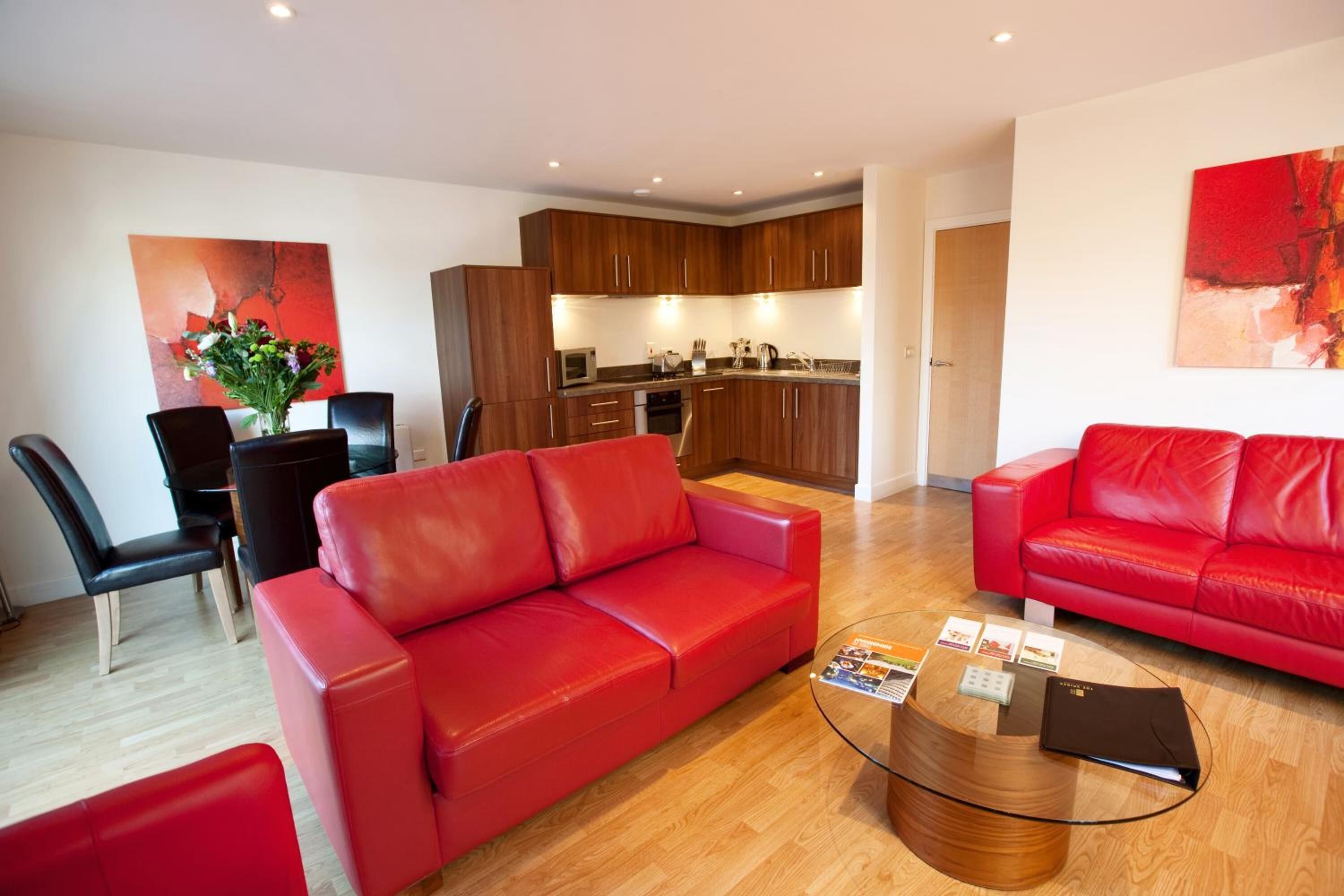 A modern living room with red leather sofas, a glass coffee table, and a kitchen in the background.