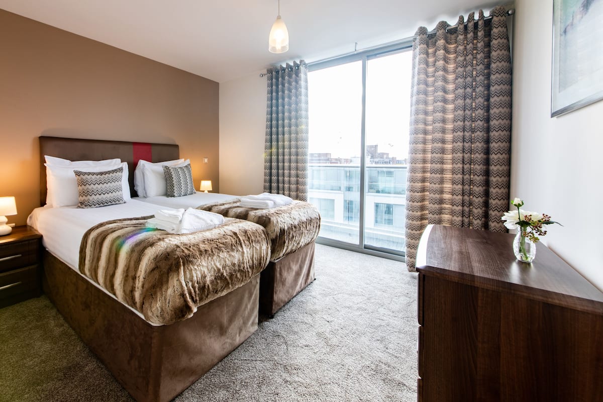 A hotel room with a large bed, a wooden nightstand with a lamp, a wooden dresser, and a view of the city through large windows.
