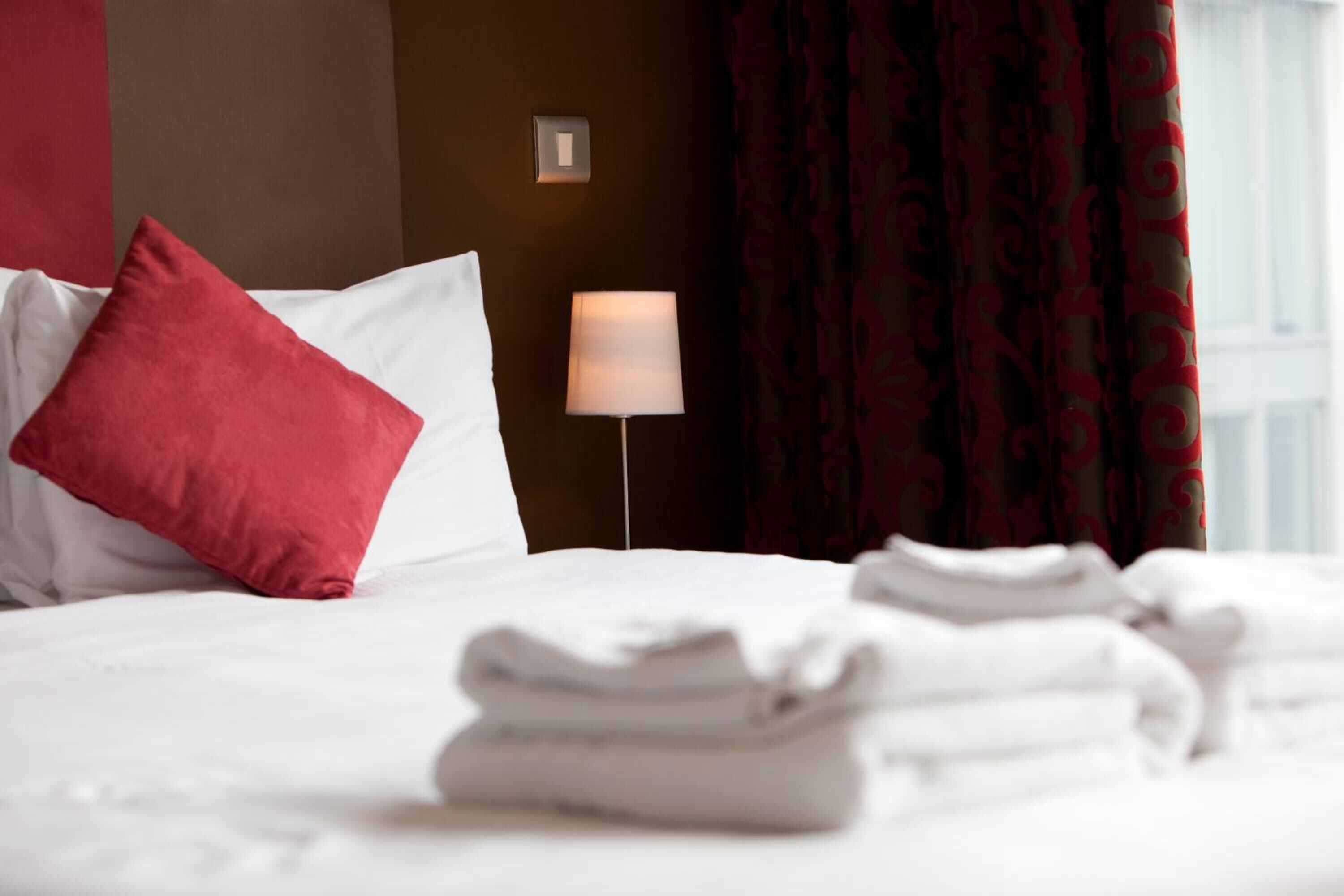 Una cama bien hecha con ropa blanca y un cojín rojo, una lámpara de noche y una ventana con cortinas estampadas.
