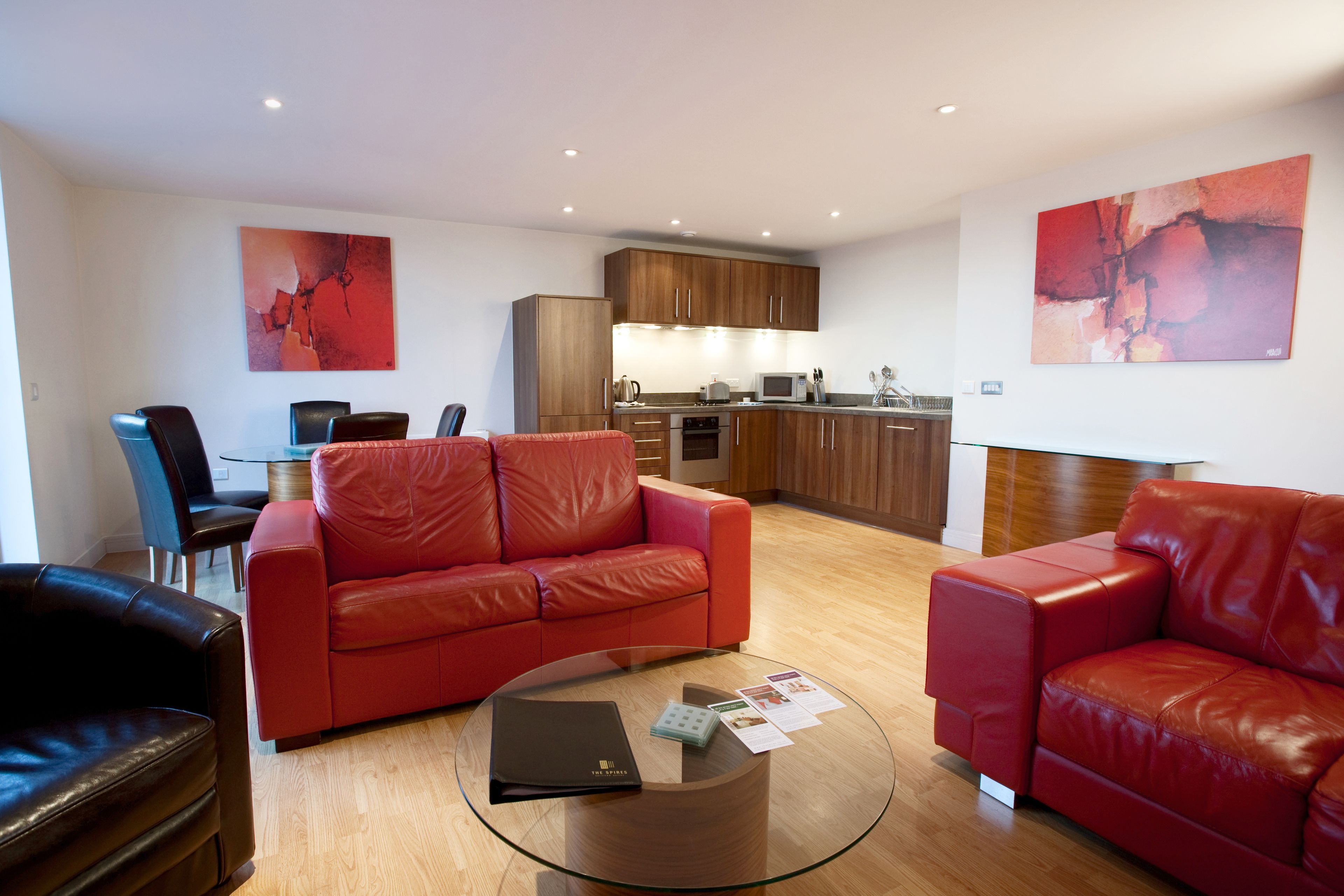 A modern living room with a red leather sofa, a glass coffee table, and a dining area with a black leather chair. A kitchen with wooden cabinets and a microwave is visible in the background.