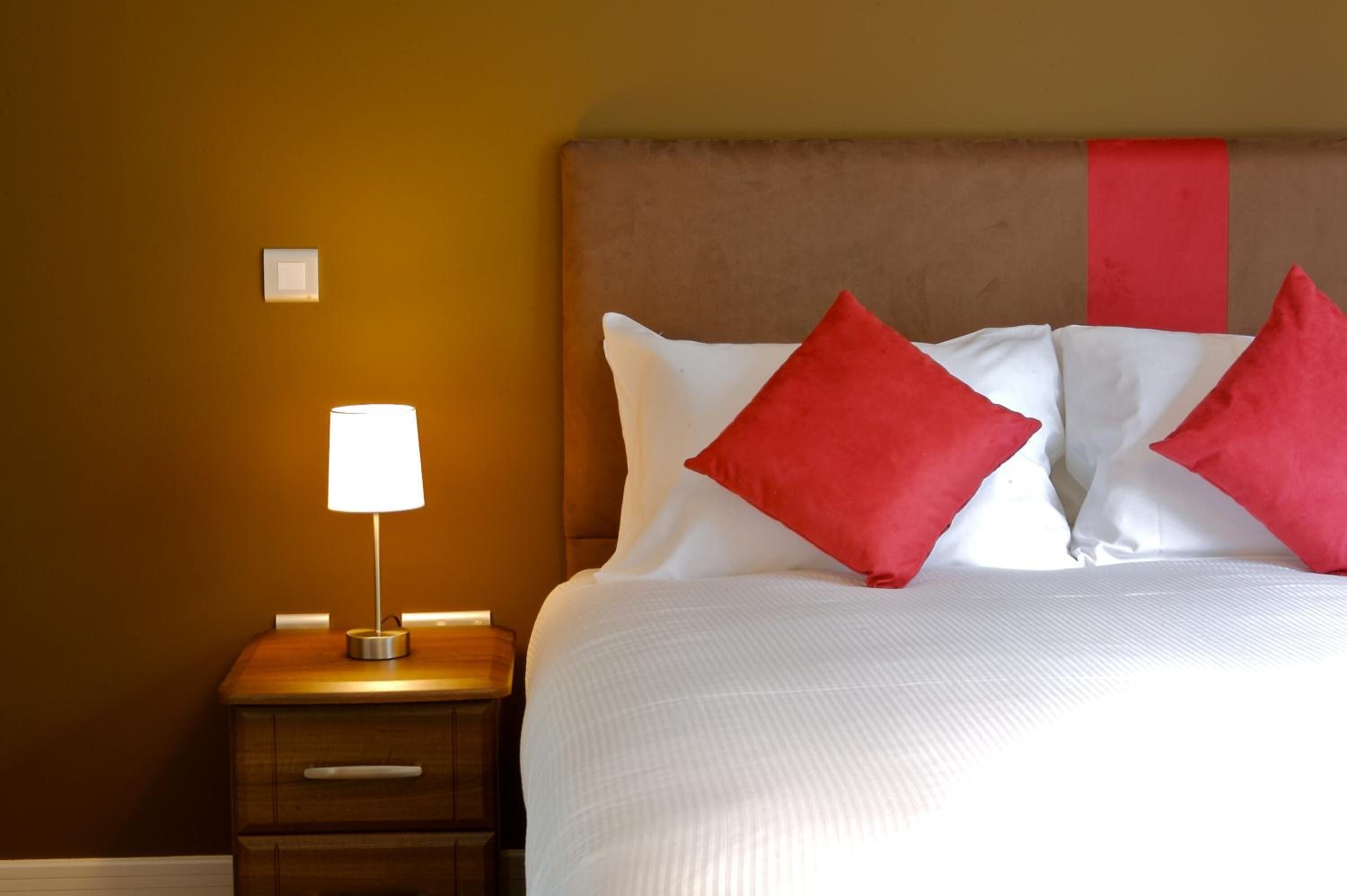 A neatly made bed with white and red pillows, a wooden nightstand with a lamp, and a wall switch.