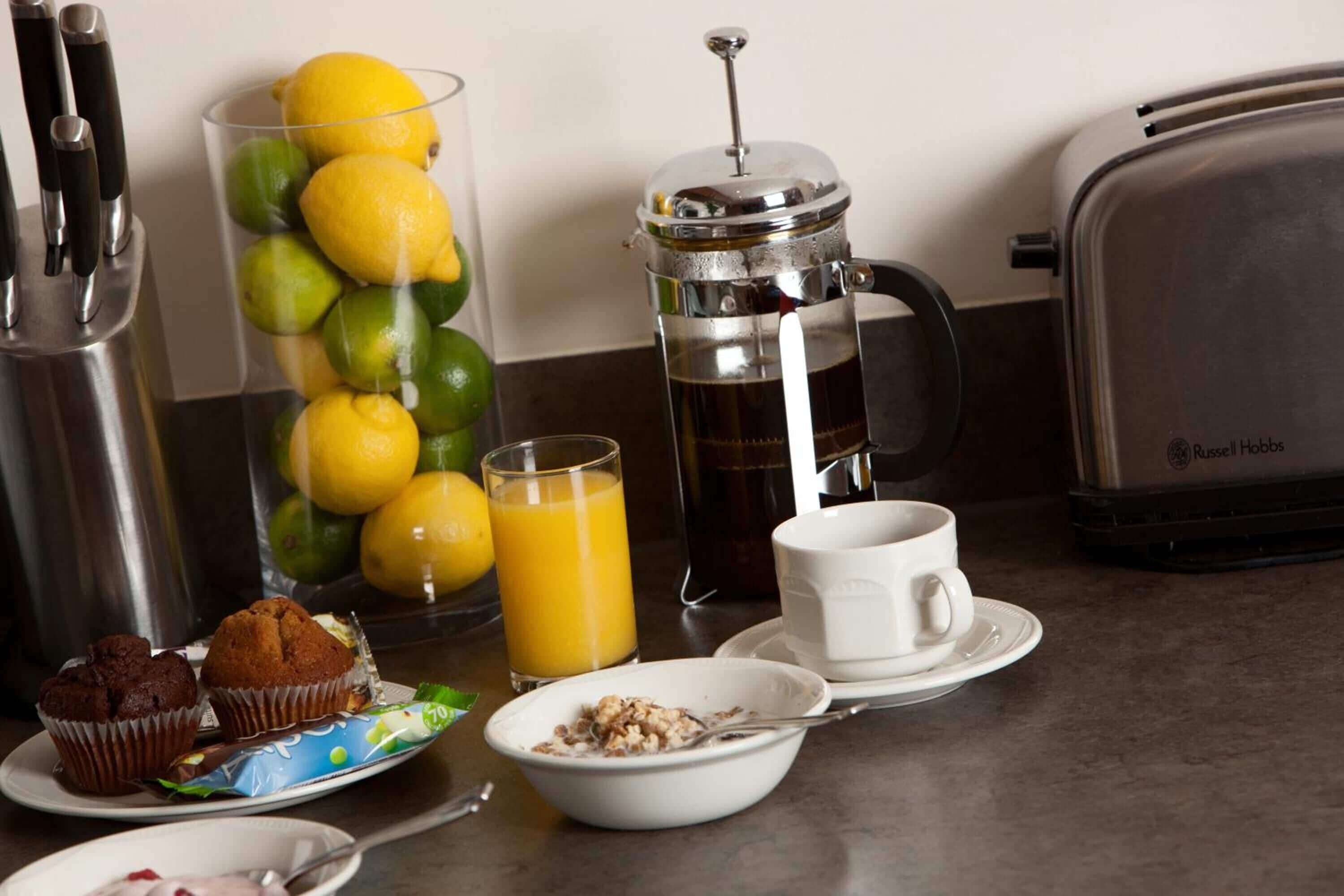A breakfast setup with a coffee press, a glass of orange juice, muffins, and a bowl of cereal.