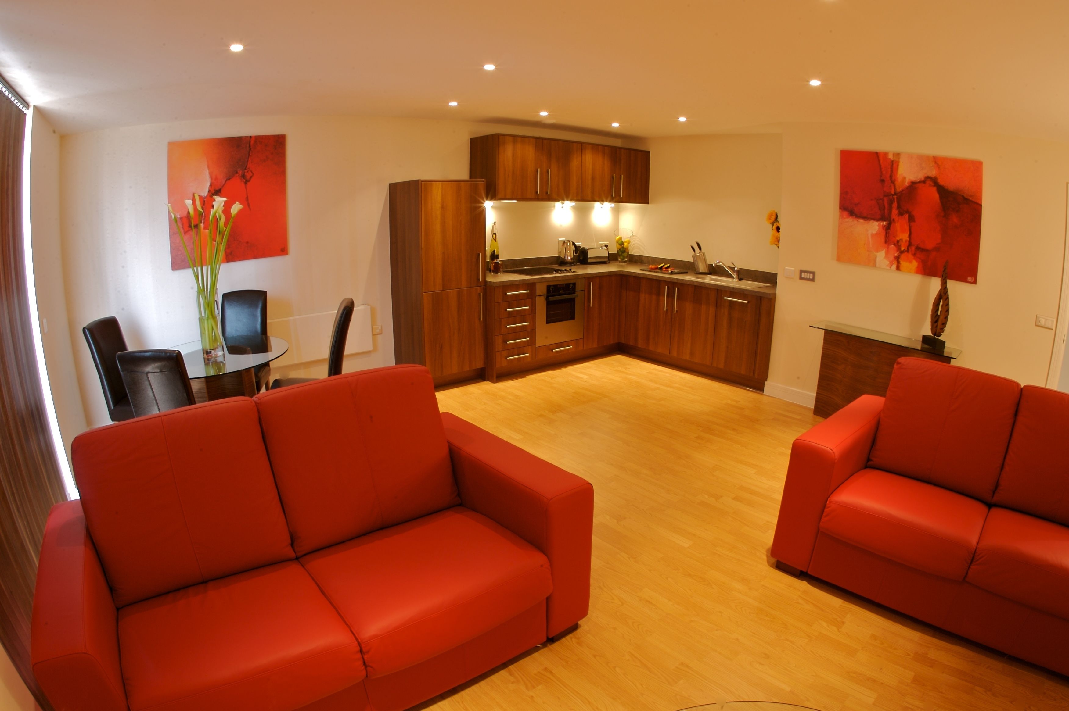 A modern living room with red sofas, a glass dining table, and a kitchen area with wooden cabinets.