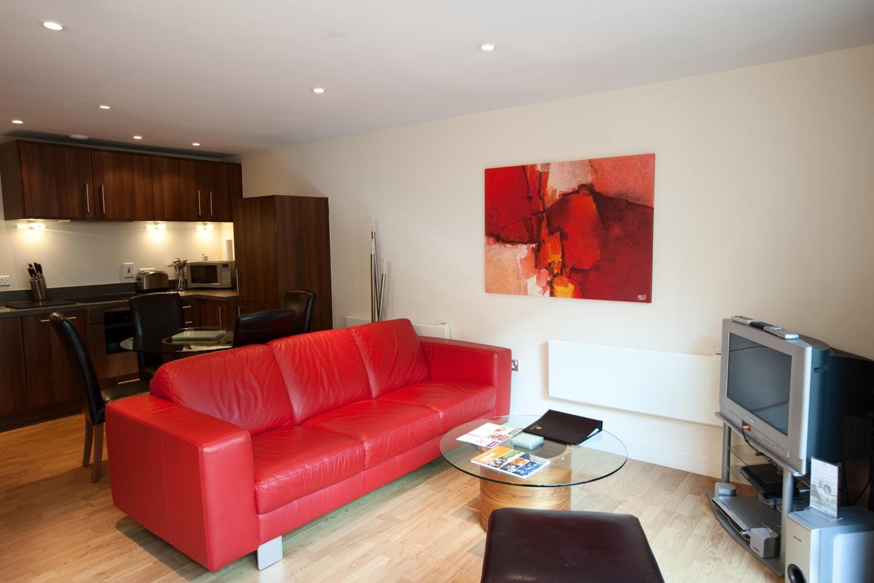 A modern living room with a red sofa, a glass coffee table, a TV, and a kitchen area with wooden cabinets and appliances.