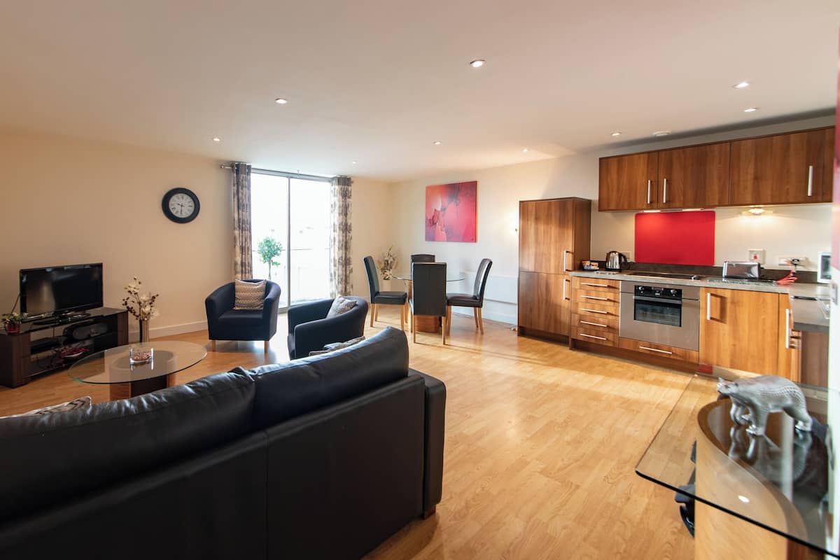 A modern living room with a sofa, a coffee table, and a TV. There is a dining area with chairs and a table. The kitchen has wooden cabinets and modern appliances.