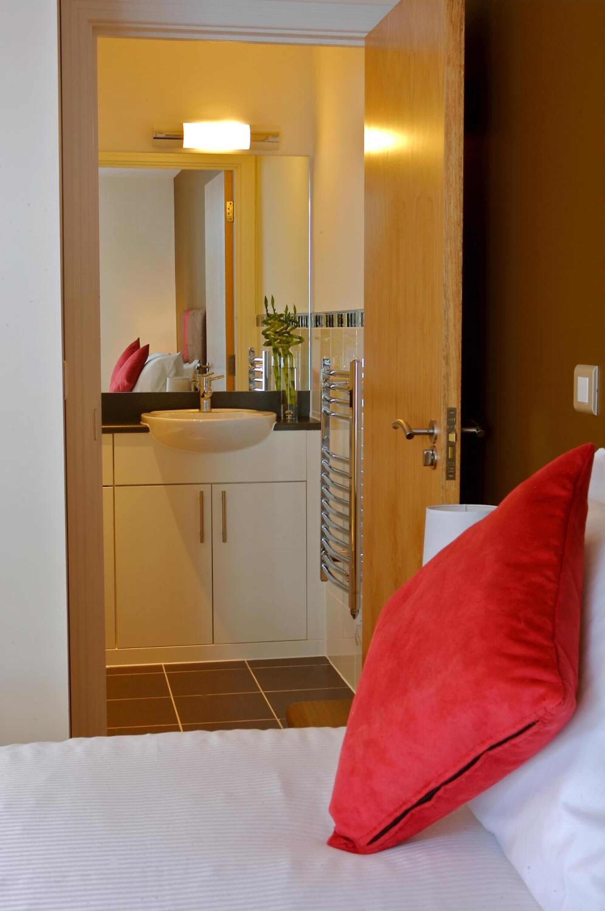 A hotel room with a bed, a red pillow, a white pillow, a wooden door, a bathroom with a sink, mirror, towel rack, and a shower.