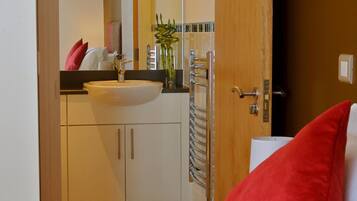 Hotelkamer met een bed, een rood kussen, een wit kussen, een houten deur, een badkamer met een wastafel, spiegel, handdoekhouder en een douche.