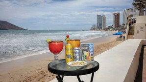 Een tafel met drankjes en hapjes op een strand, met uitzicht op de oceaan en gebouwen op de achtergrond.