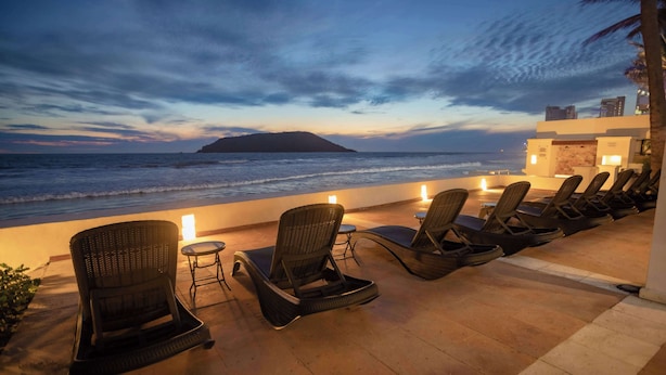Een strandlounge met rieten stoelen en tafels, met uitzicht op de oceaan en een eiland in de verte.