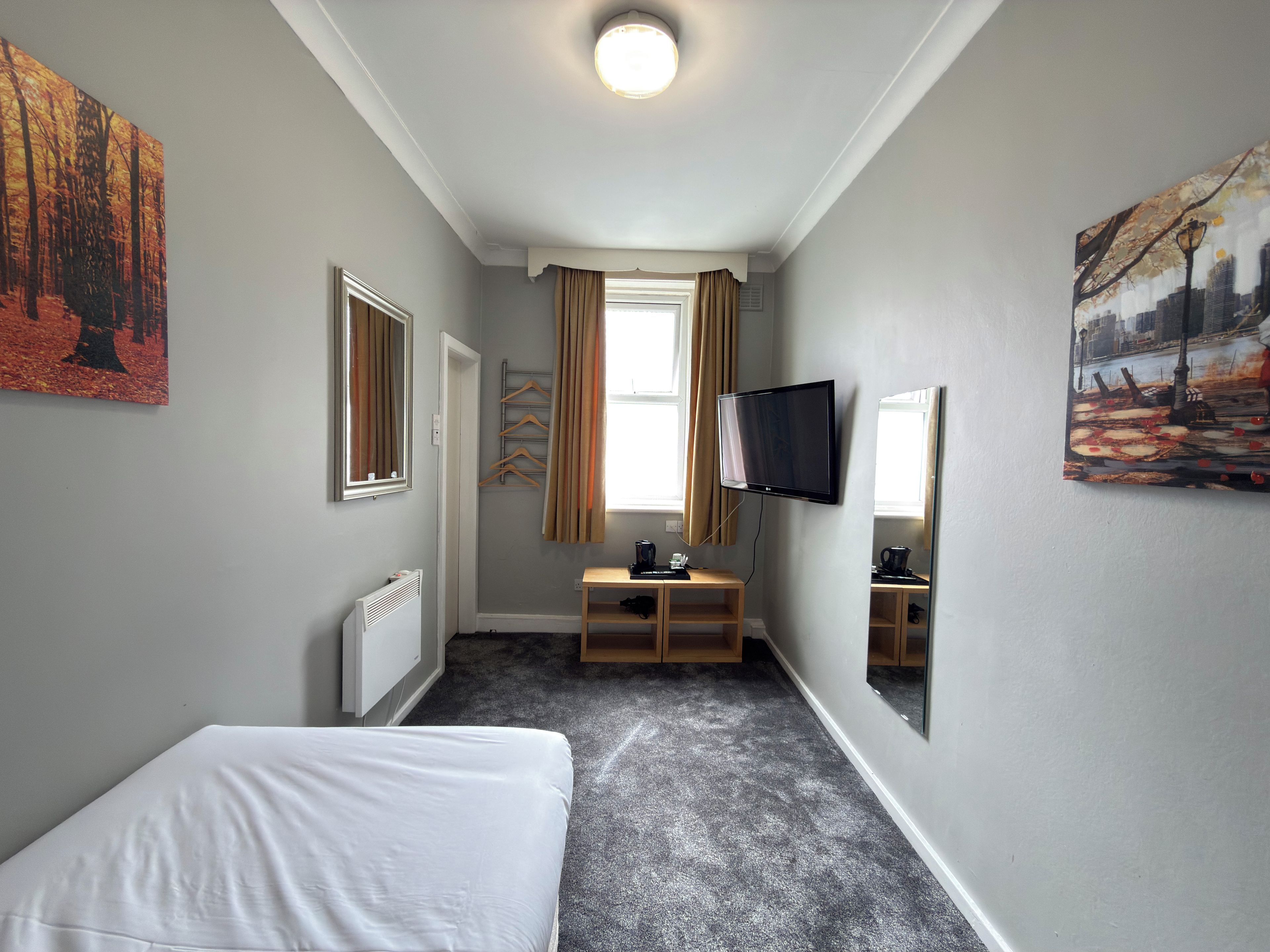A hotel room with a bed, a TV, a radiator, a small table with a coffee maker, and a window with curtains.