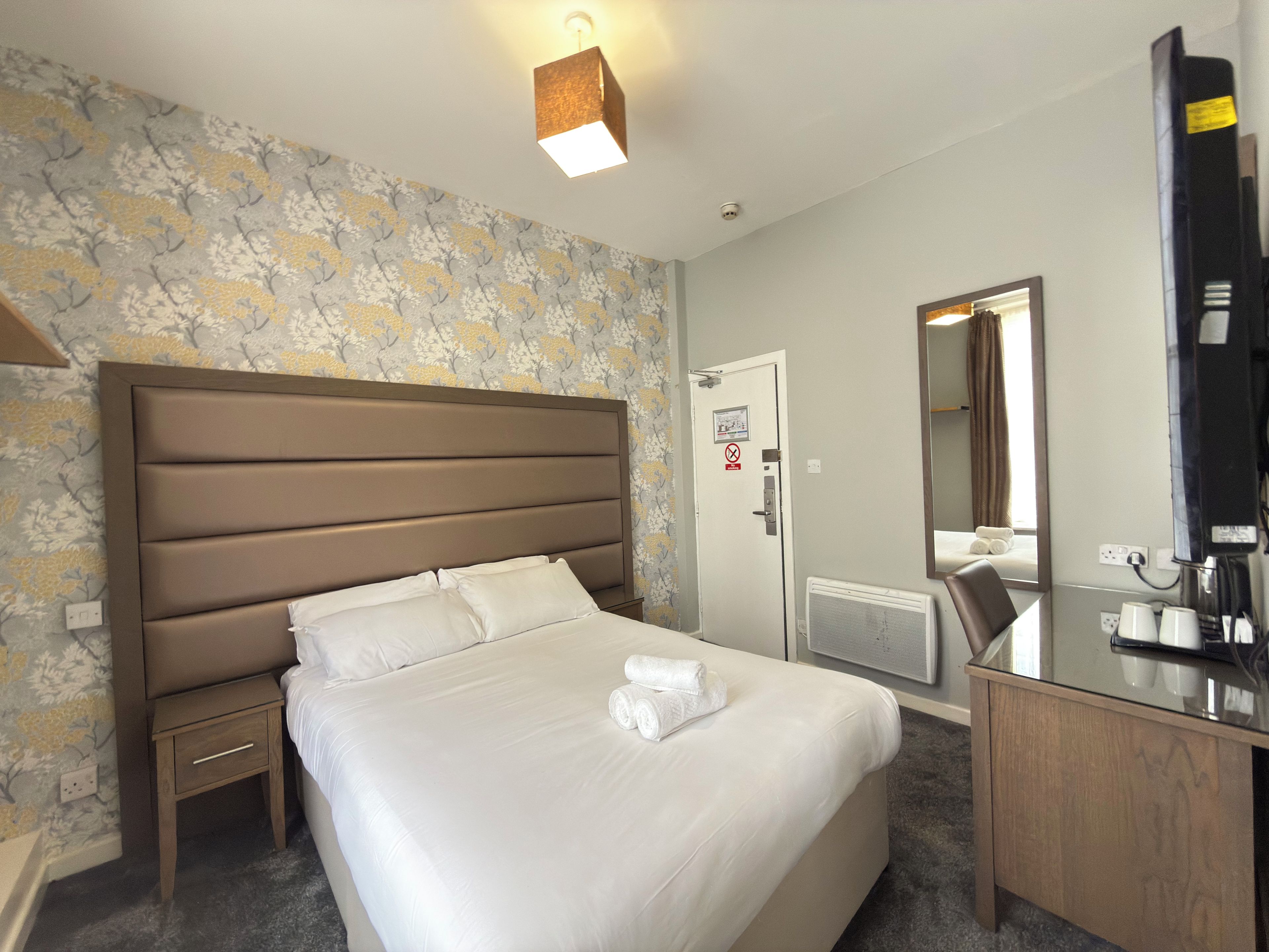 Ein Hotelzimmer mit einem großen Bett, einem Nachttisch, einem Schreibtisch mit Spiegel und einem an der Wand befestigten Fernseher.