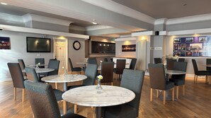 Un espace repas moderne avec des tables rondes en marbre et des chaises rembourrées, un téléviseur à écran plat fixé au mur et une horloge.