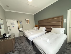 Ein Hotelzimmer mit zwei Einzelbetten, einem Schreibtisch mit Stuhl, einem Spiegel und einer Tür mit Handtuchhalter.