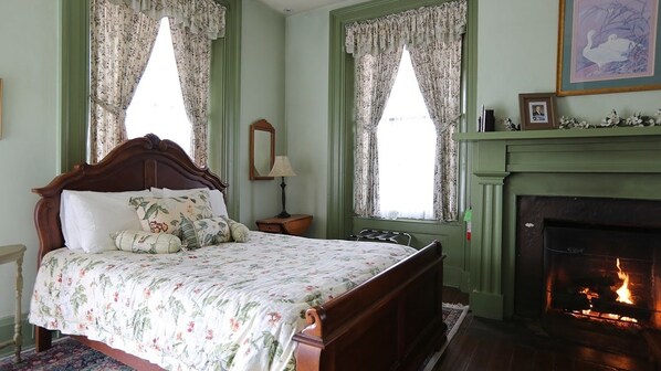 A bedroom with a wooden bed, floral bedding, a fireplace, and two windows with curtains.
