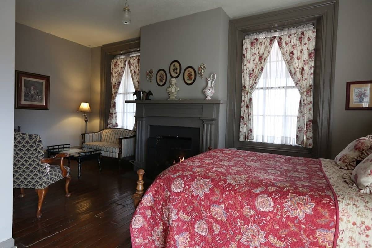 A room with a fireplace, a bed with a floral bedspread, a patterned armchair, and a lamp.