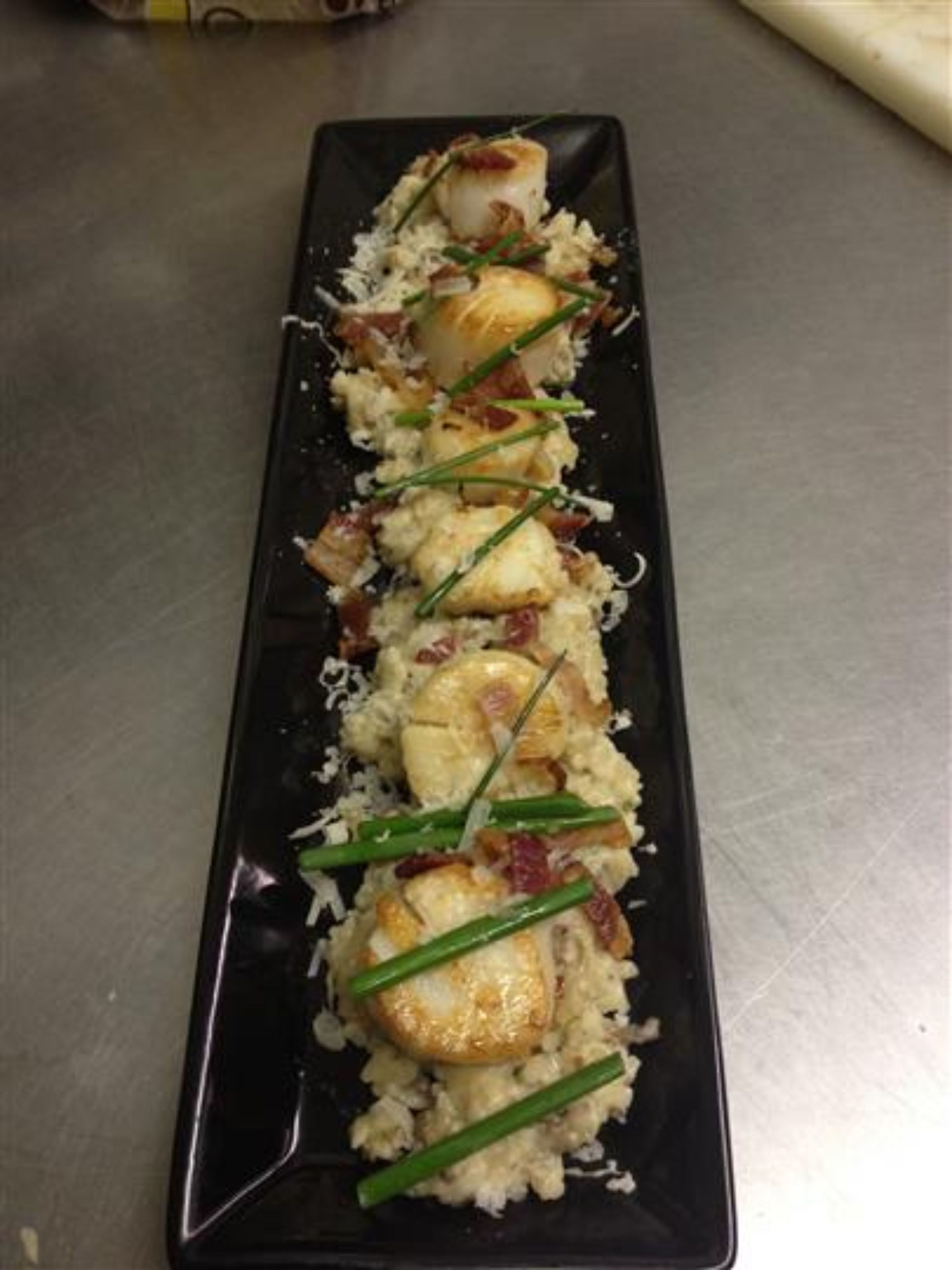 A black rectangular plate with a food arrangement consisting of rice, green onions, and pieces of food that appear to be chicken or fish, possibly in a sushi or similar dish.