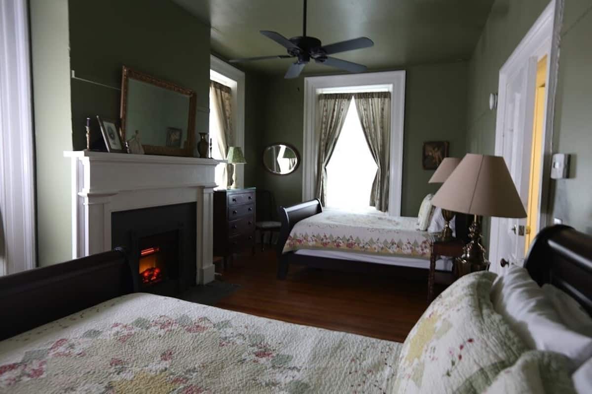 A bedroom with two beds, a fireplace, a ceiling fan, and a large window with curtains.
