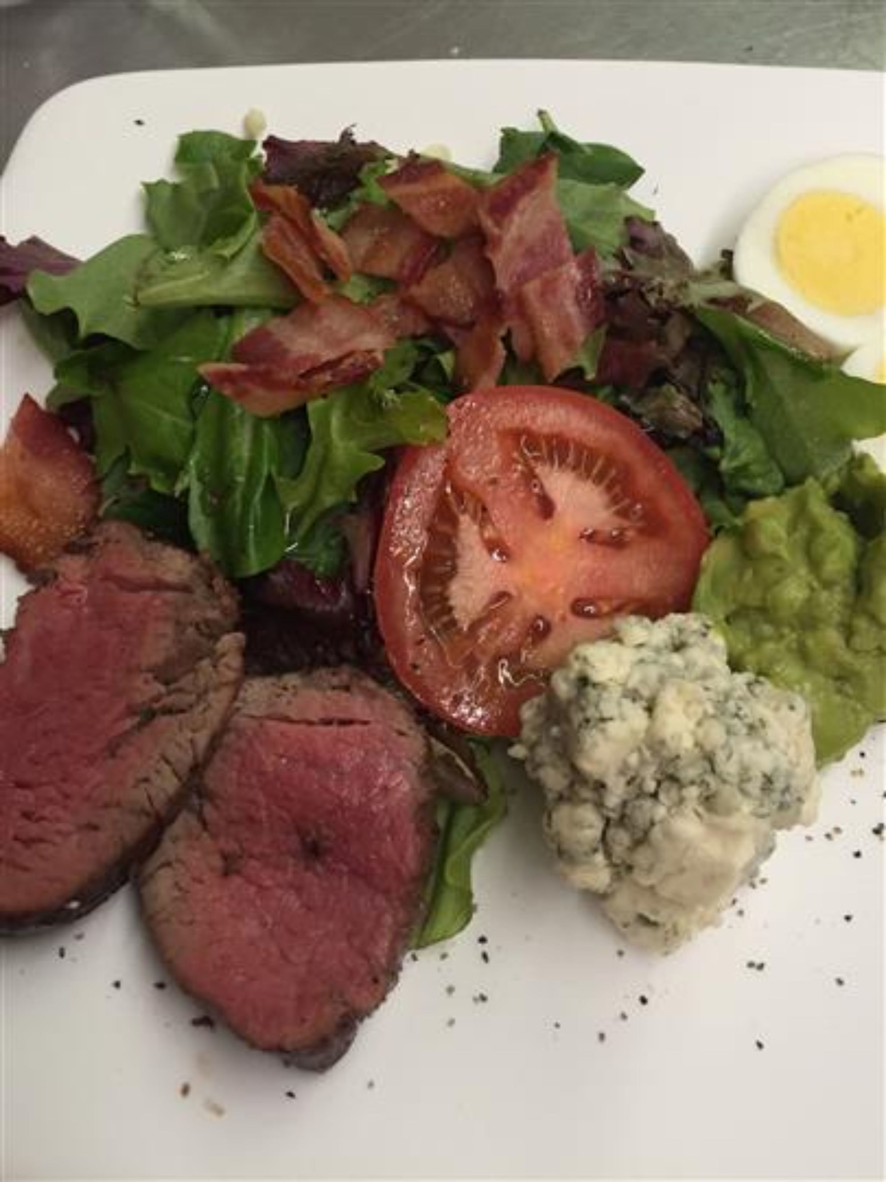 A plate with a salad, a slice of tomato, a piece of meat, a boiled egg, and a dollop of blue cheese.
