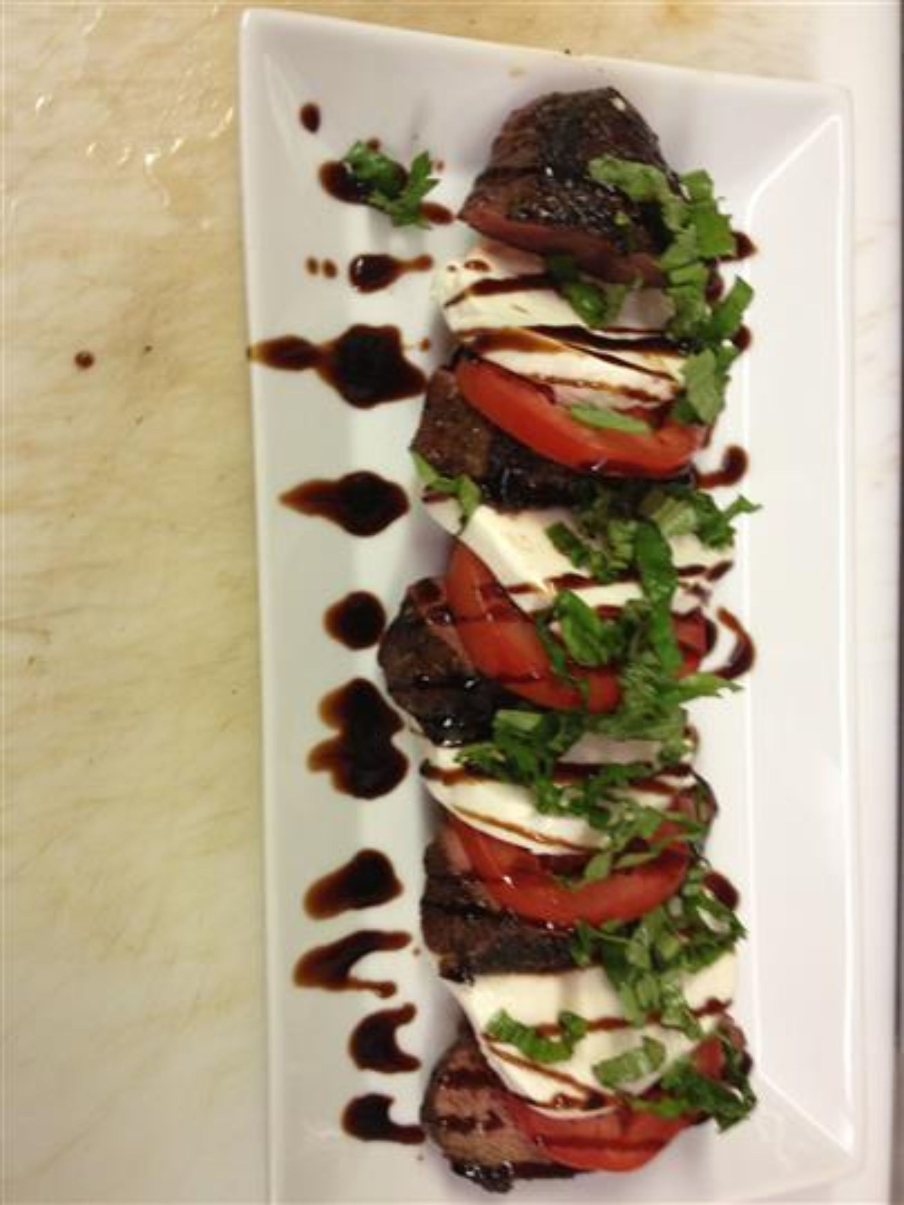 A rectangular plate with a food arrangement consisting of grilled meat, tomato slices, and a white creamy topping, garnished with green herbs, and drizzled with a dark sauce.