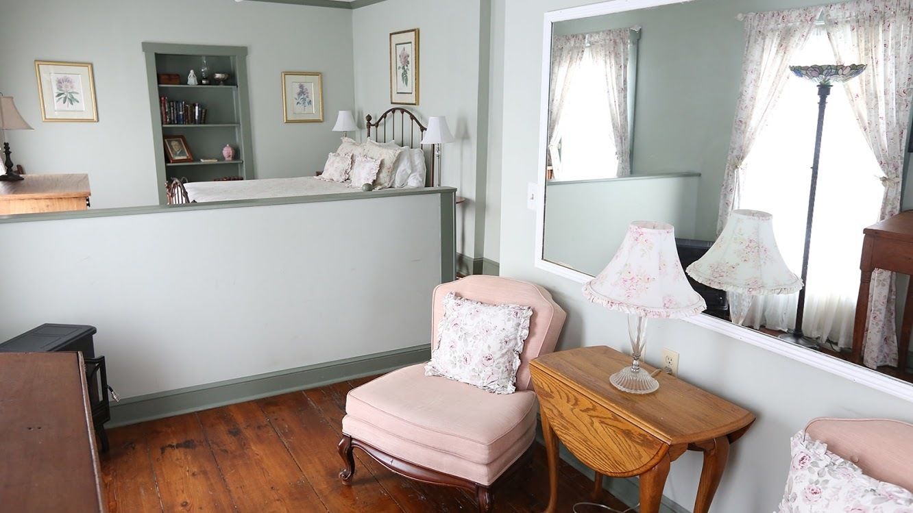 Un dormitorio con cama, una silla, una mesa, un espejo y una estantería con libros y objetos decorativos.