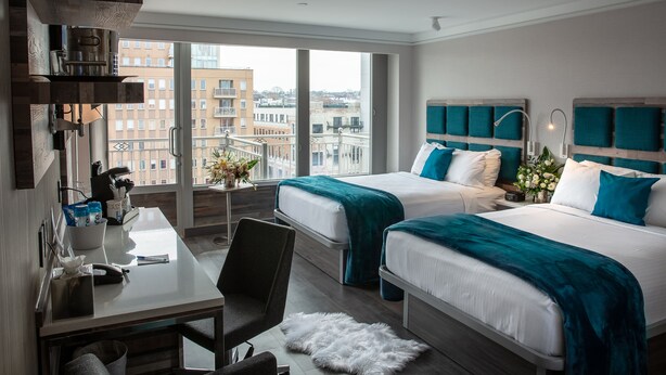 A hotel room with two beds, a desk, a chair, and a view of the city. - Hotel Le Bleu (Brooklyn)