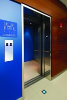 Egy lift, melynek a falán kék szín van, és a jobb oldalon egy fa borítás látható. A falon egy tábla található, amelyen a vészhelyzeti utasítások szerepelnek, a lift ajtajai felett pedig egy vészhelyzeti kijáratra utaló tábla.