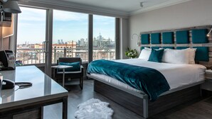 A modern hotel room with a large bed, a desk, and a view of the city skyline. - Hotel Le Bleu (Brooklyn)