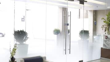 Un área de recepción moderna con puerta de vidrio, sofá blanco, alfombra azul y plantas en macetas.