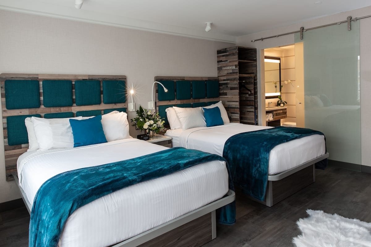 Un dormitorio moderno con dos camas, una mesita de noche, un espejo y un baño visible a través de una puerta corrediza.