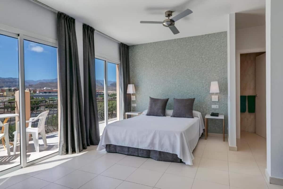 Une chambre d’hôtel moderne dotée d’un grand lit, de deux tables de chevet, d’un ventilateur de plafond et offrant une vue sur la ville grâce à de grandes fenêtres du sol au plafond.