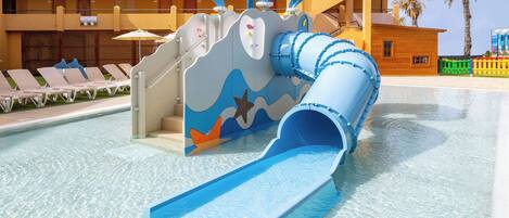 A water slide with a blue and white design, a pool, and a building with balconies in the background.