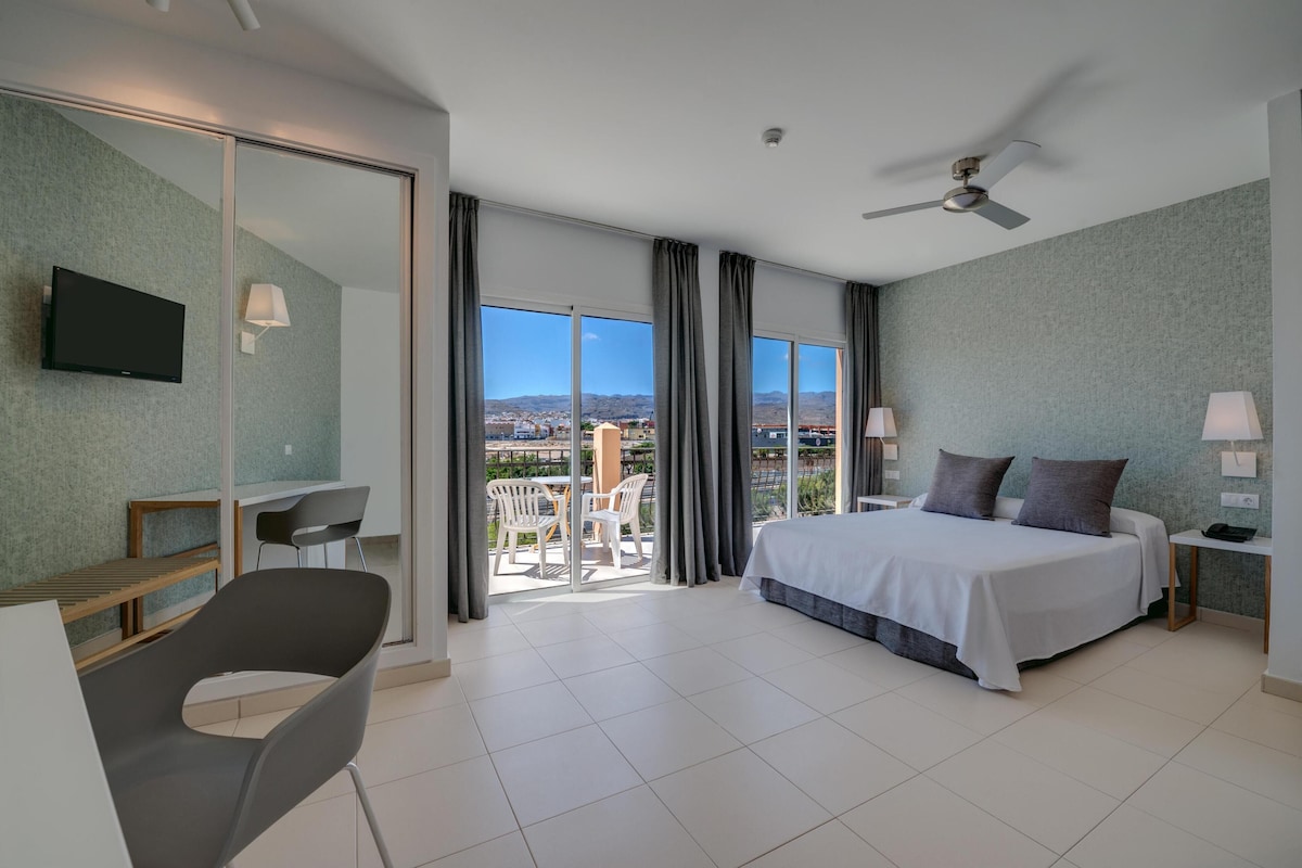 Una camera d'albergo moderna con un letto grande, una scrivania, una sedia e un balcone con vista sulla città e sulle montagne.