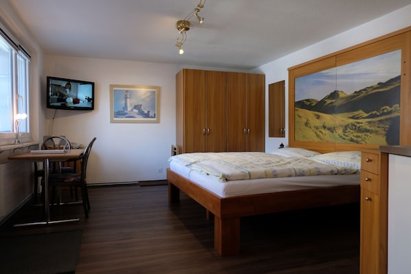 A bedroom with a wooden bed, a desk with a chair, a television, and a painting on the wall. - Artist Apartments & Hotel Garni (Zermatt)
