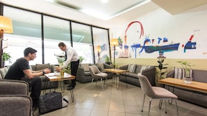 Un moderno hall de hotel con diversas opciones de asientos, incluyendo sofás y mesas, un gran mural abstracto en la pared y vistas amplias a través de grandes ventanales.