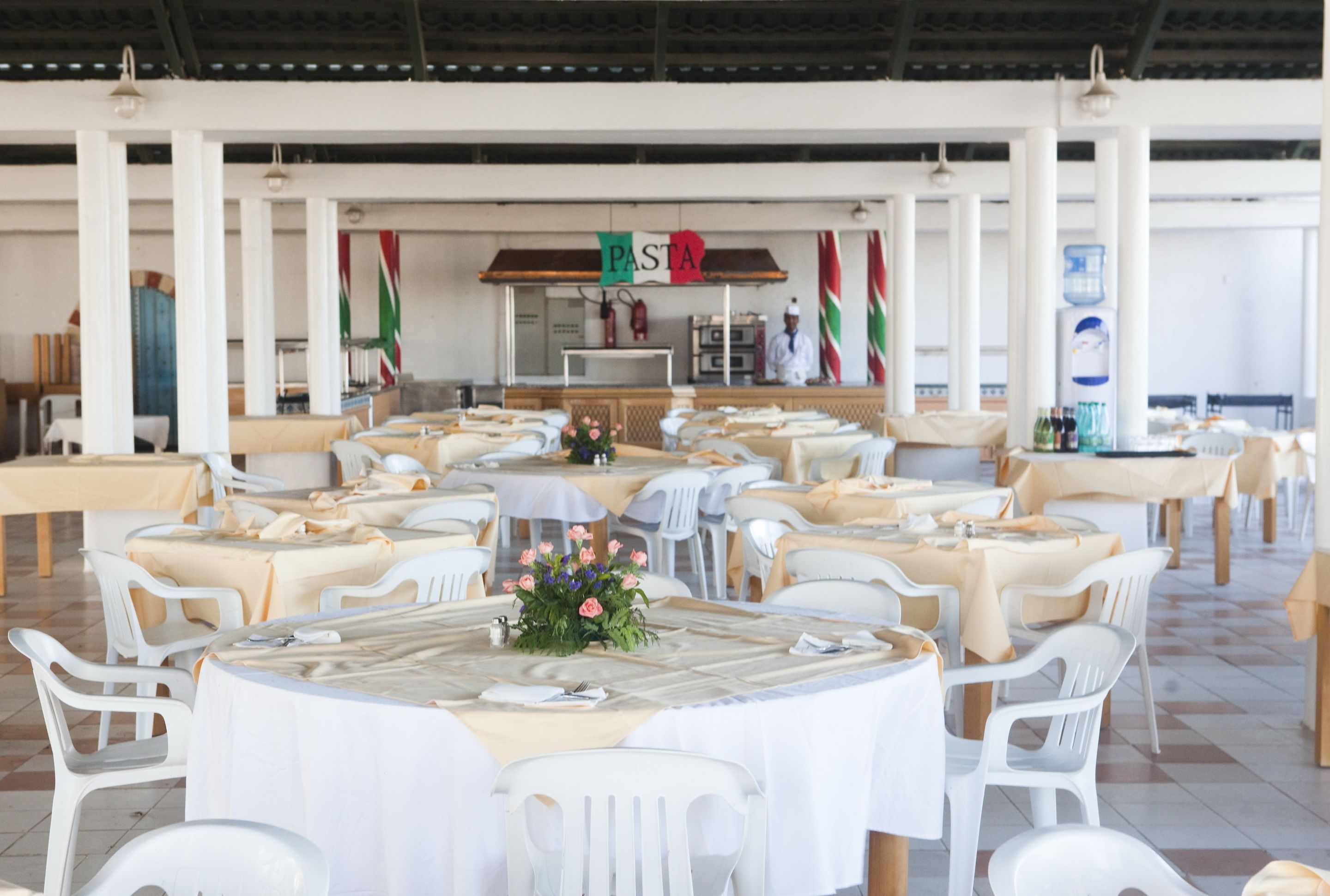 A banquet hall with round tables and white chairs, a pasta station, and a buffet area.